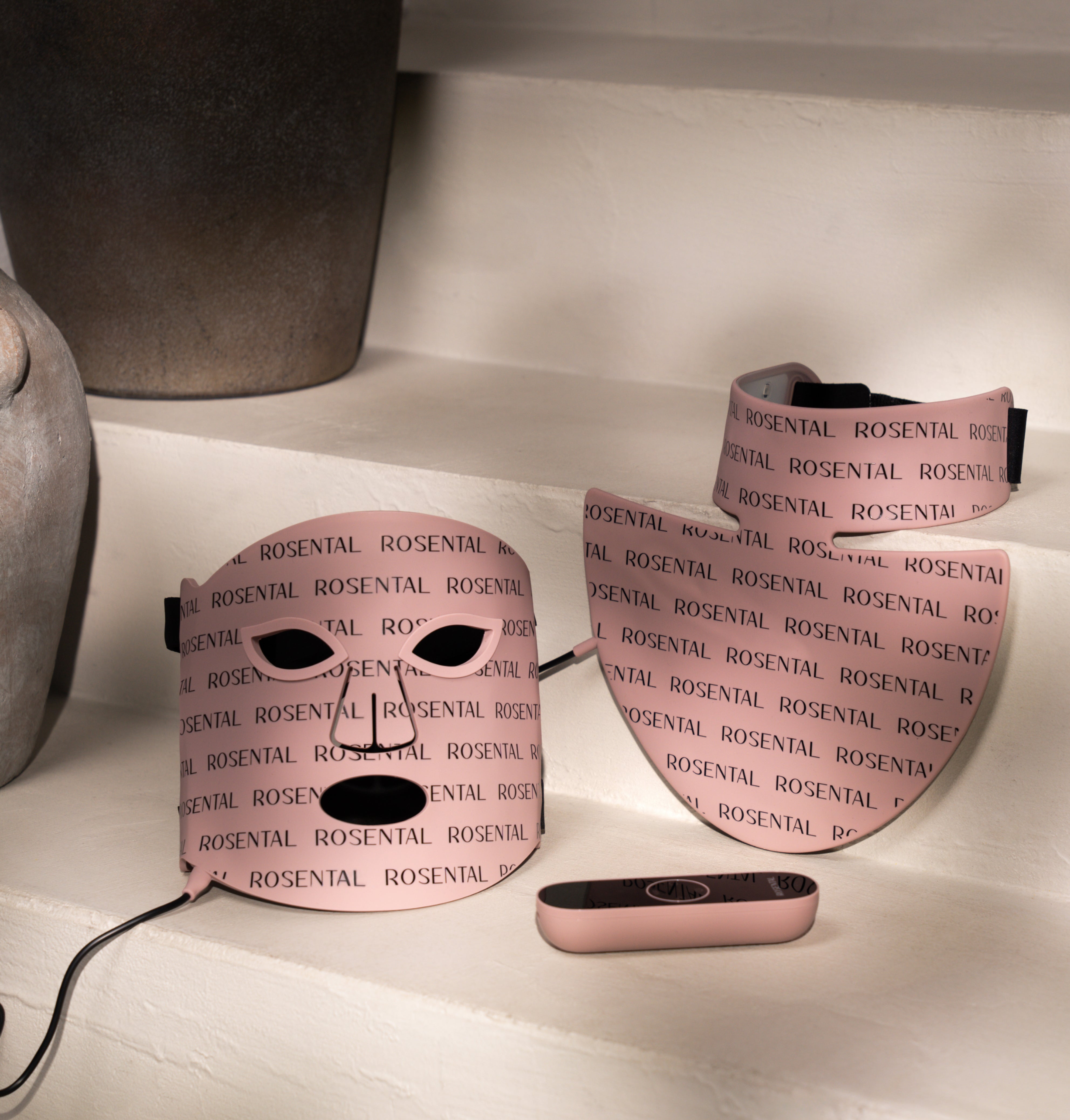 Lichttherapiemaskers met het "ROSENTAL"-logo liggen decoratief op een witte plank naast stenen vaten.