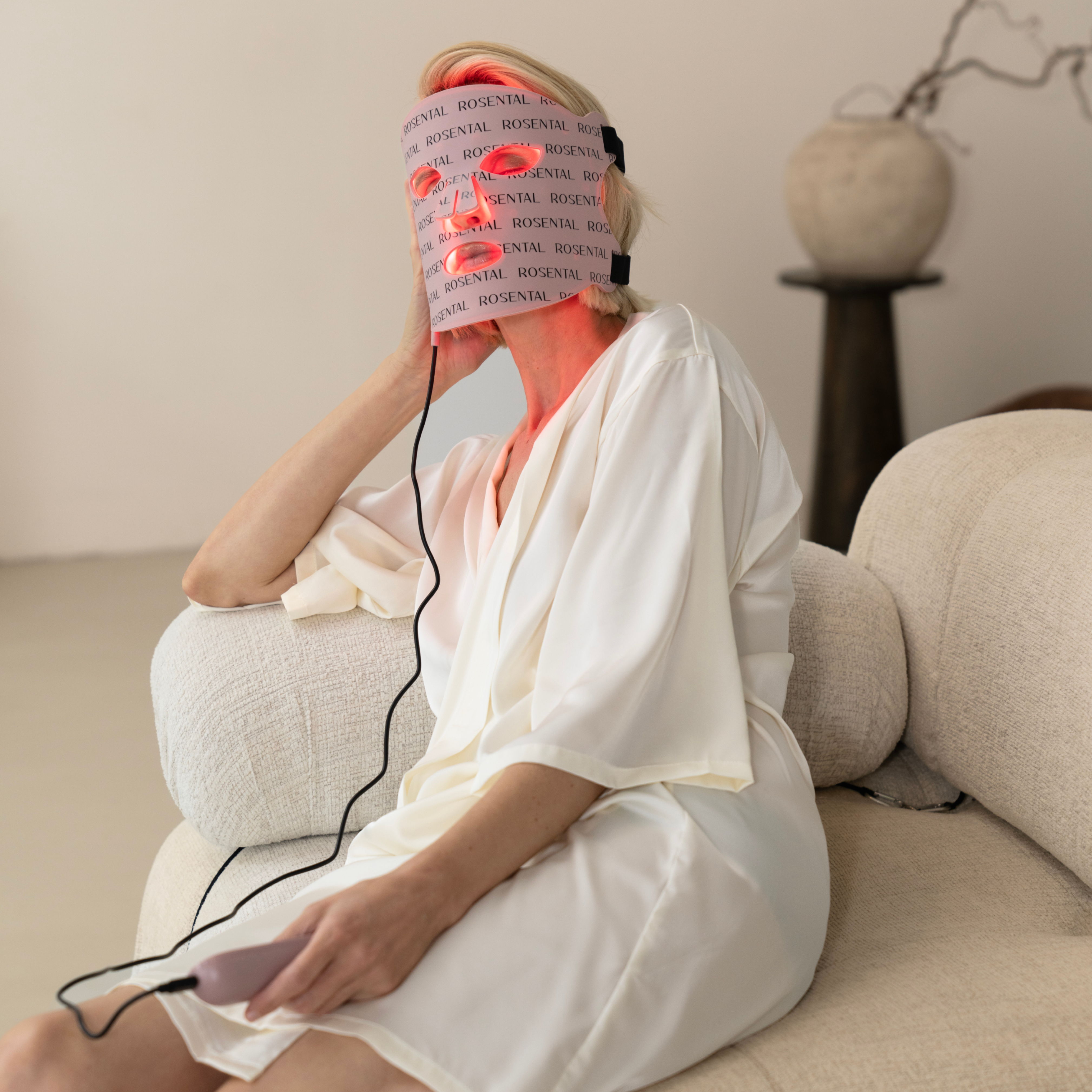 Een vrouw in een badjas zit op de bank en gebruikt het LED-gezichtsmasker van Rosental Organics.